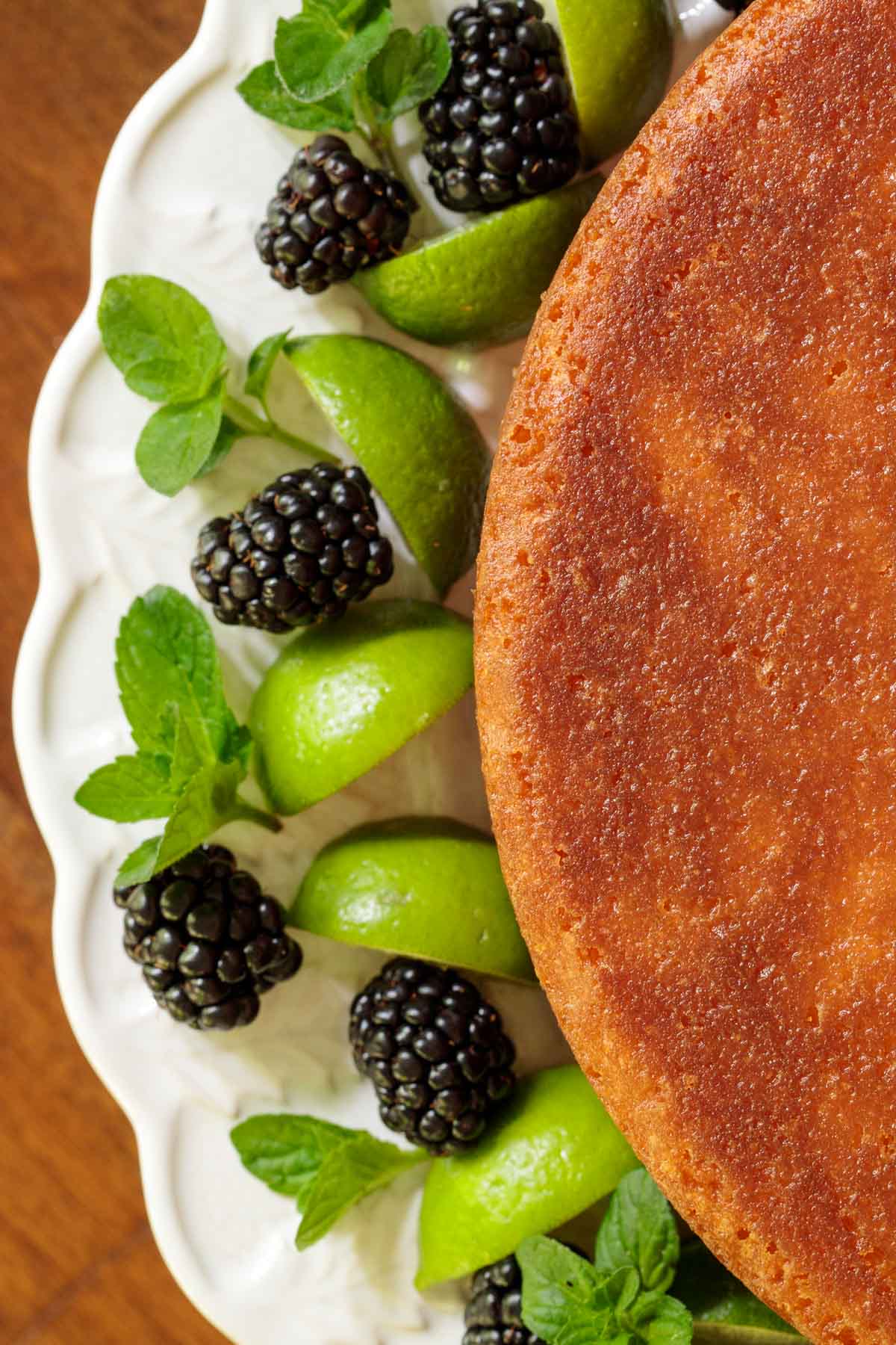 Overhead closeup photo of an Easy Key Lime Coconut Cake surrounded by blackberries, mint leaves and lime wedges on a white cake stand.