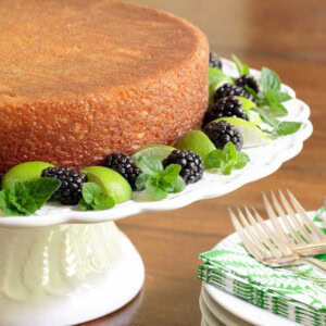 Vertical picture of key lime crunch cake on a white cake stand garnished with key limes, mint and blackberries
