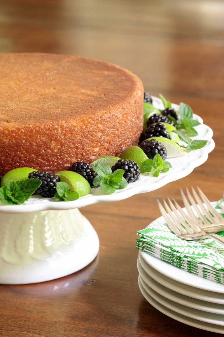 Vertical picture of key lime crunch cake on a white cake stand garnished with key limes, mint and blackberries