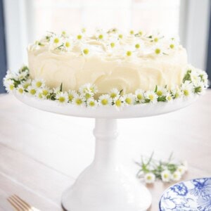 Vertical picture of lemon curd poppy seed cake decorated with flowers on a white cake stand.