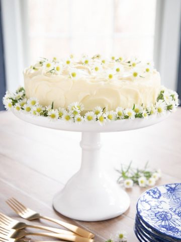 Vertical picture of lemon curd poppy seed cake decorated with flowers on a white cake stand.