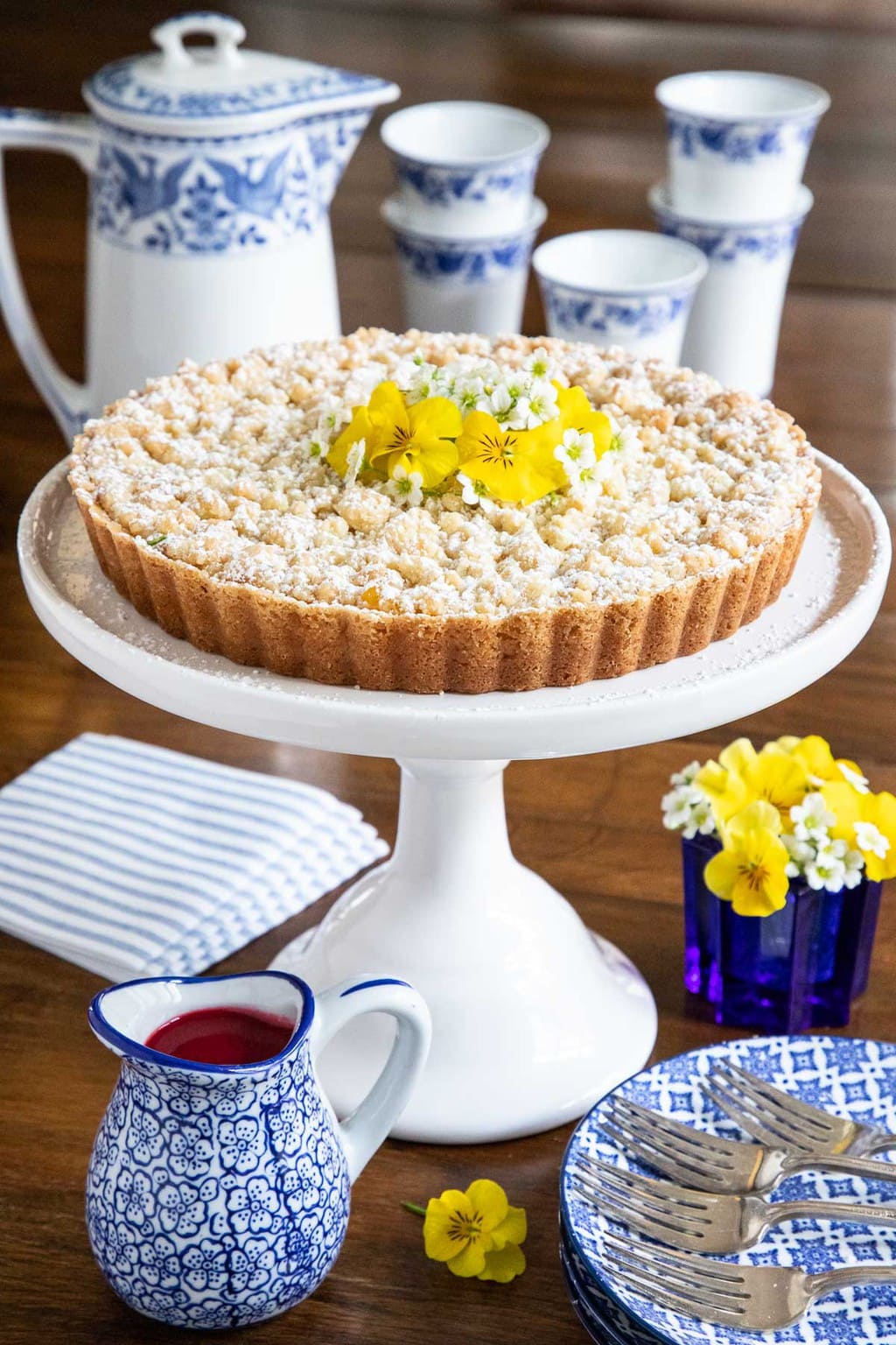Vertical photo of a Lemon Curd Shortbread Tart on a white pedestal cake stand with a jar of Raspberry Coulee in the foreground.