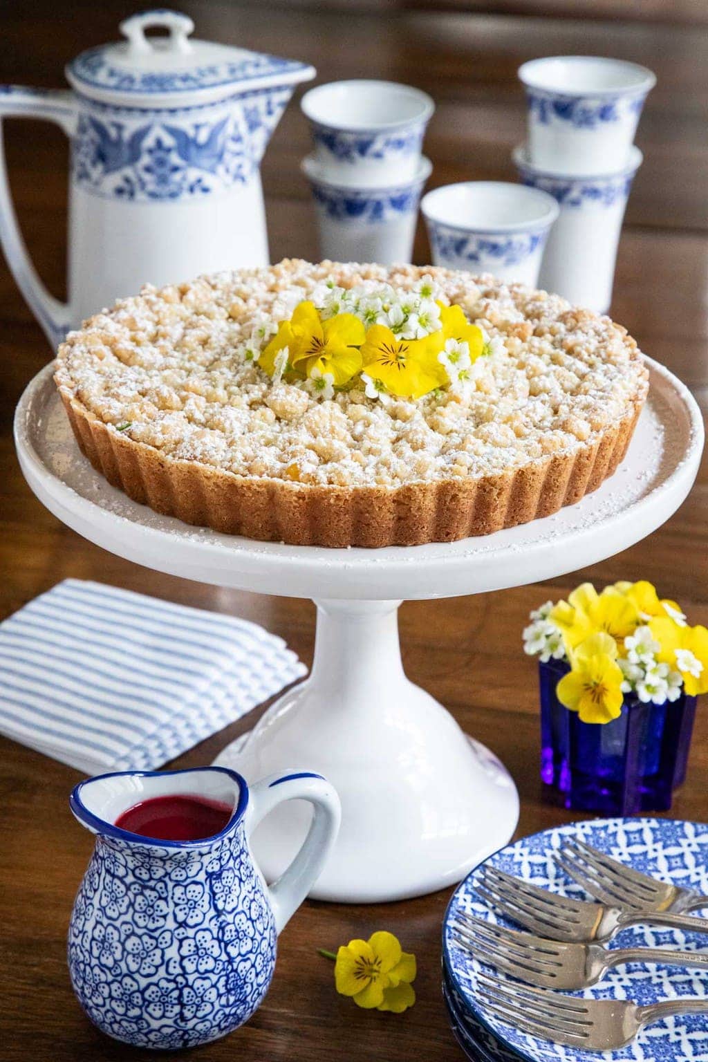 Vertical photo of Lemon Curd Shortbread Tart on a white pedestal cake stand decorated with yellow pansies.