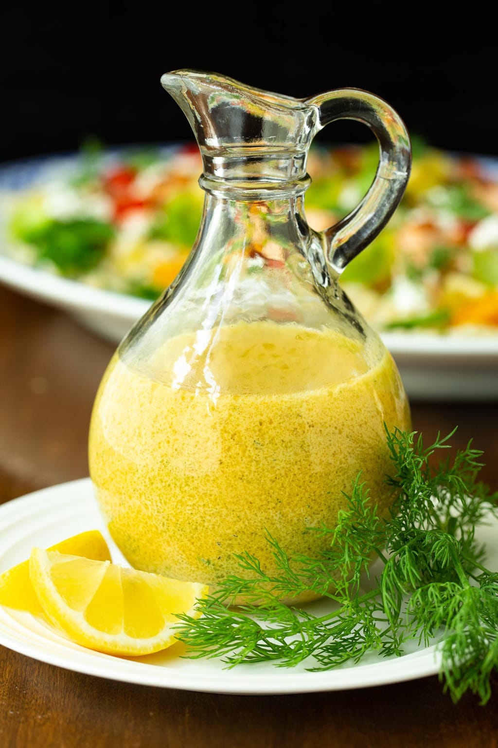 Vertical closeup photo of Lemon Dill Dressing in a glass cruet with fresh lemons and dill.