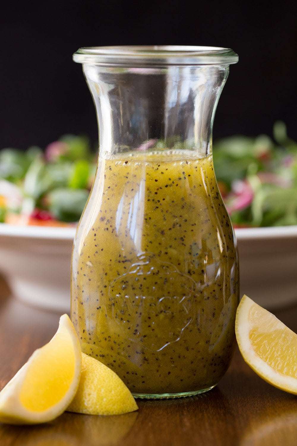 Vertical photo of Lemon Ginger Poppyseed Salad Dressing in a glass cruet with fresh lemon wedges in the foreground and a green salad in the background.