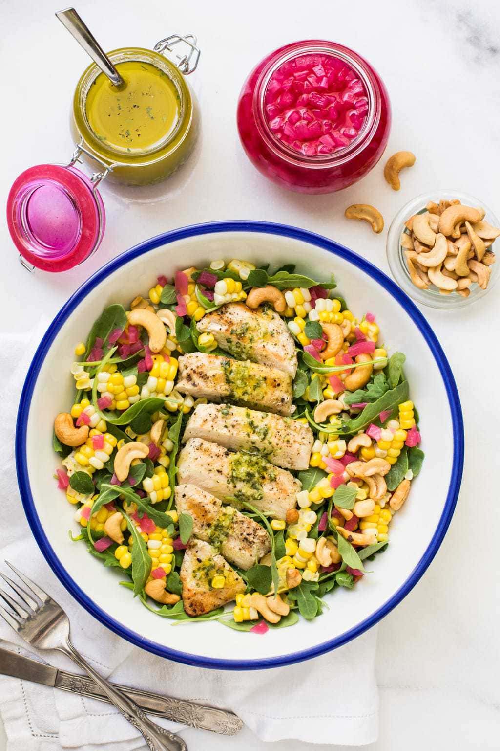 Overhead photo of a serving bowl filled with Lemon Oregano Chicken and Corn Salad. Surrounding the bowl are many of the ingredients used in making this salad.
