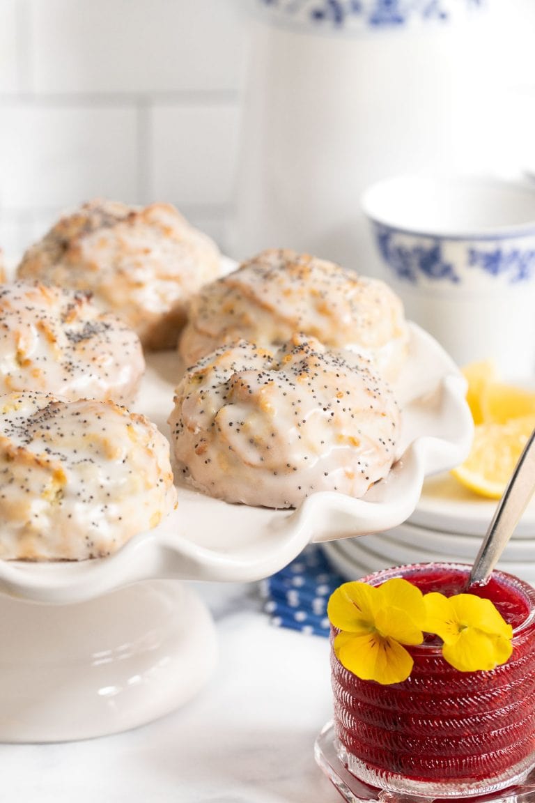 Vertical photo of a batch of Lemon Poppy Seed Scones on a white pedestal serving platter with a small jar of Raspberry Freezer Jam in the foreground.