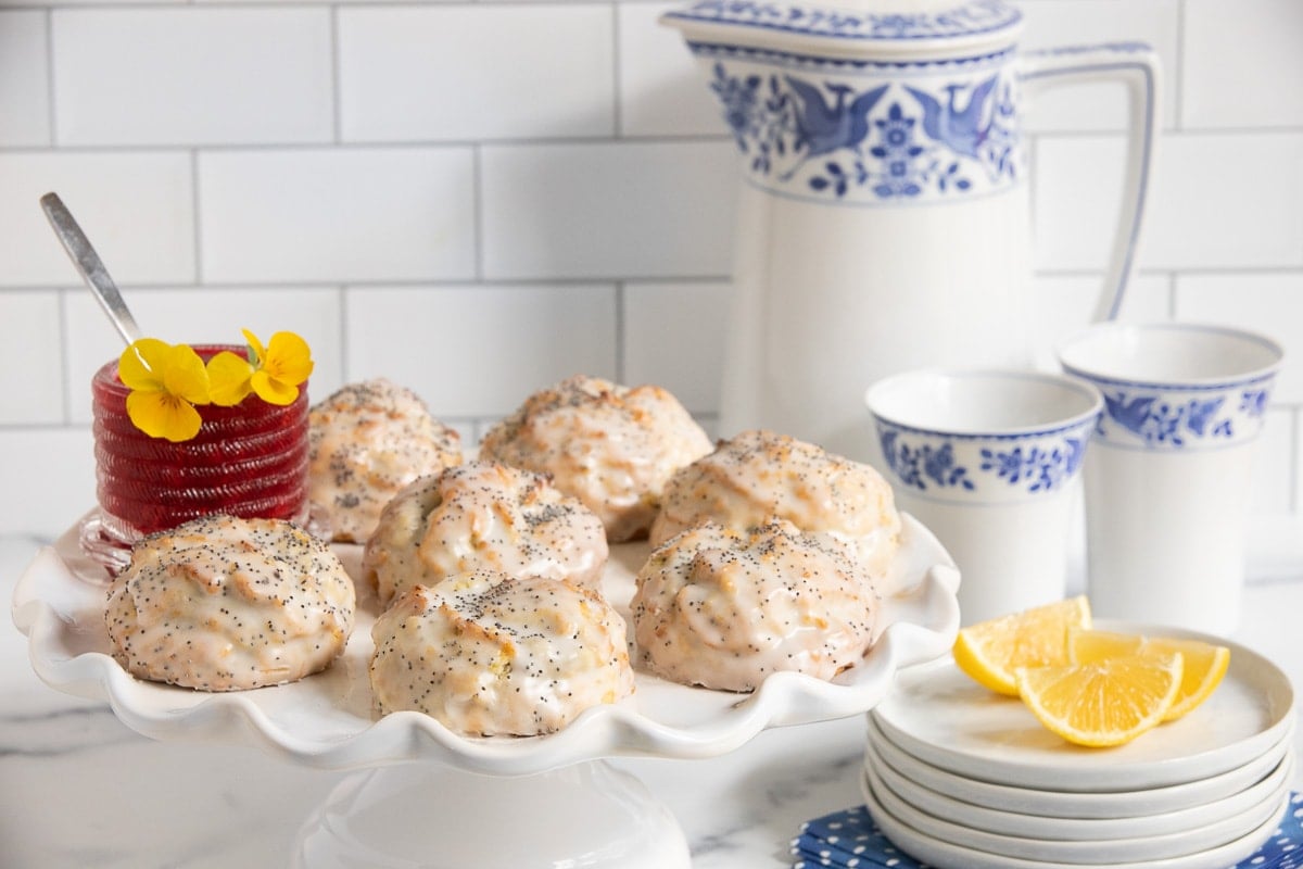 Horizontal photo of a batch of Ridiculously Easy Lemon Poppy Seed Scones on a scalloped pedestal serving plate with with a jar of Raspberry Freezer Jam.