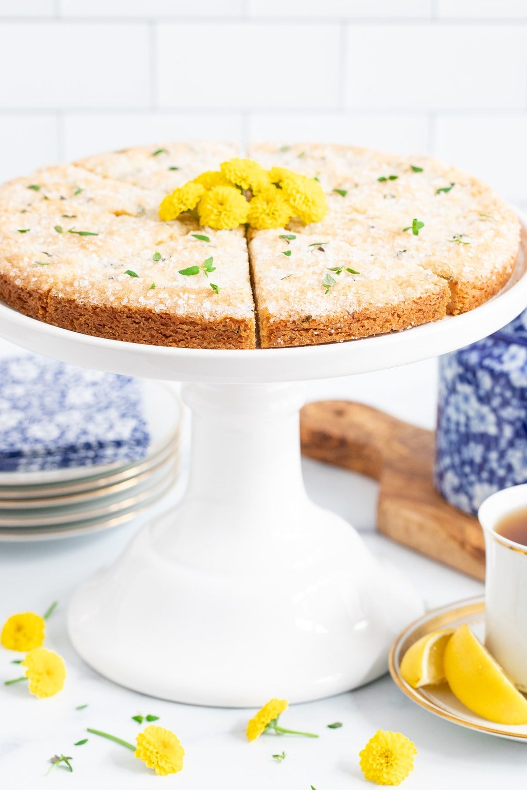 Vertical closeup photo of Lemon Thyme Shortbread on a white pedestal stand surrounded by yellow daisies, a cup of tea and dessert plates.