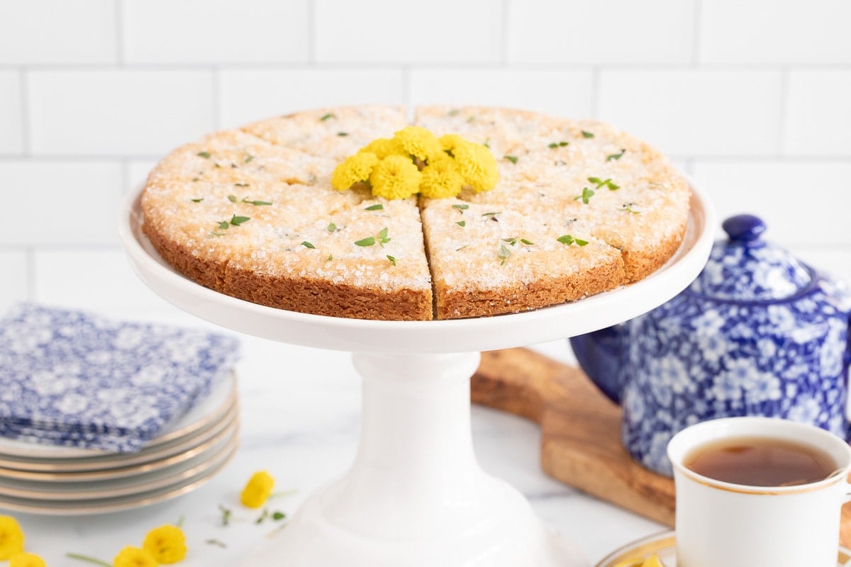 Horizontal photo of a white pedestal presentation plate filled with Lemon Thyme Shortbread garnished with fresh thyme leaves and yellow daisies.