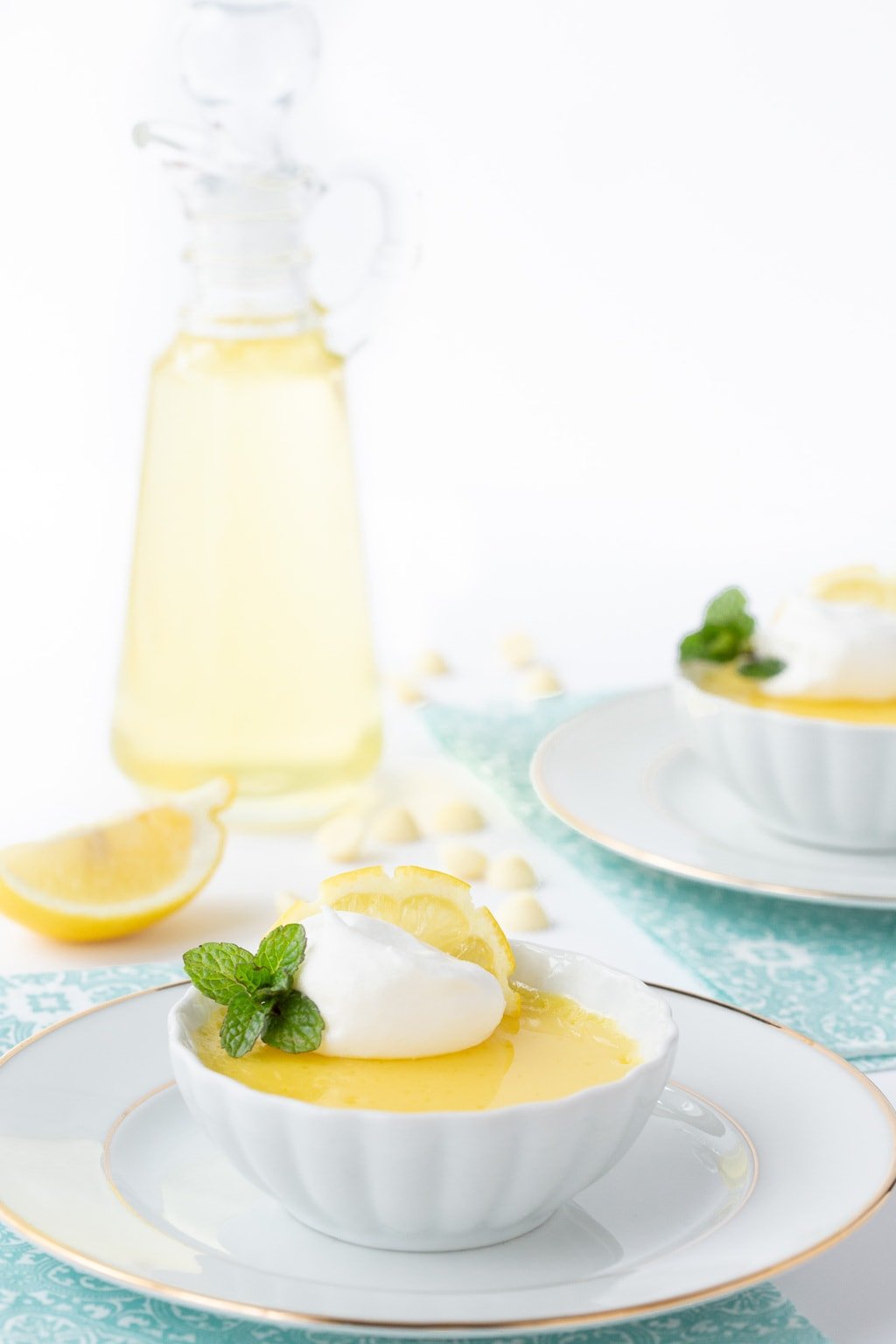 Vertical photo of Lemon and White Chocolate Pots de Crème in small white dishes with a glass pitcher of lemoncello syrup in the background.