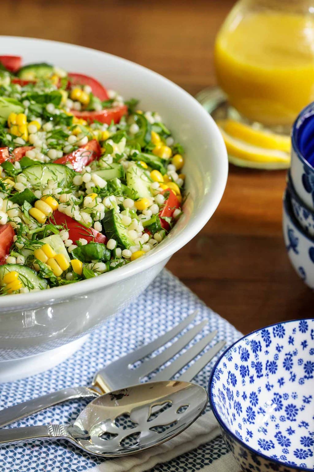 Closeup vertical photo of a Mediterranean Couscous Salad in a white serving bowl surrounded by serving accessories and a glass pitcher of lemon dressing.