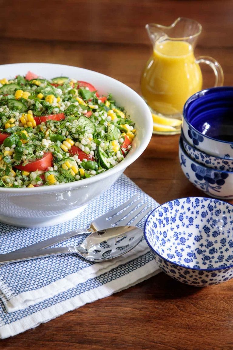 Vertical picture of lemony couscous salad in a white bowl on a wooden table with small blue and white bowls stacked