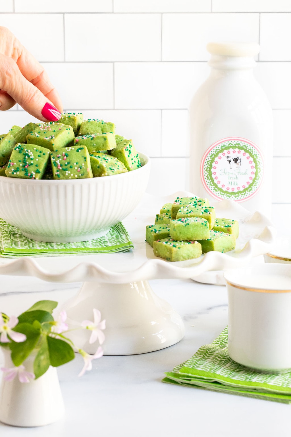 Vertical photo of a bowl of Leprechaun Shortbread Bites on a pedestal display stand with a shamrock plant in the foreground and a quart of Irish milk in the background. A hand is taking one of the shortbread bites.