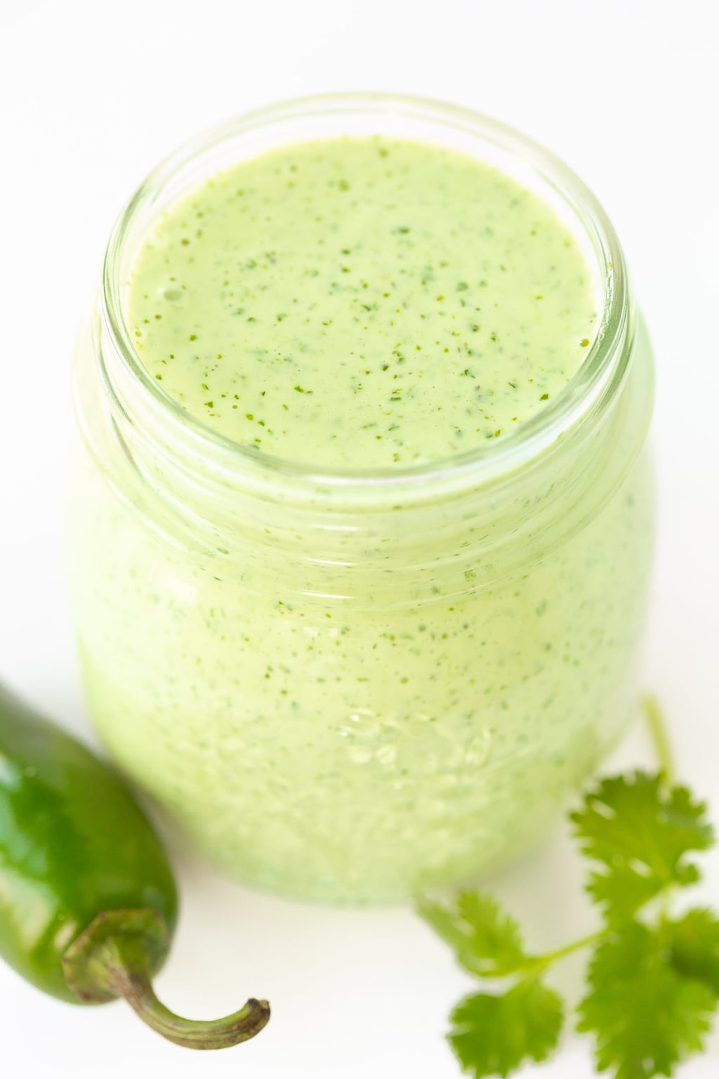 Vertical overhead photo of Light and Spicy Cilantro Dressing in a glass jar surrounded by Jalepeno peppers and cilantro leaves.