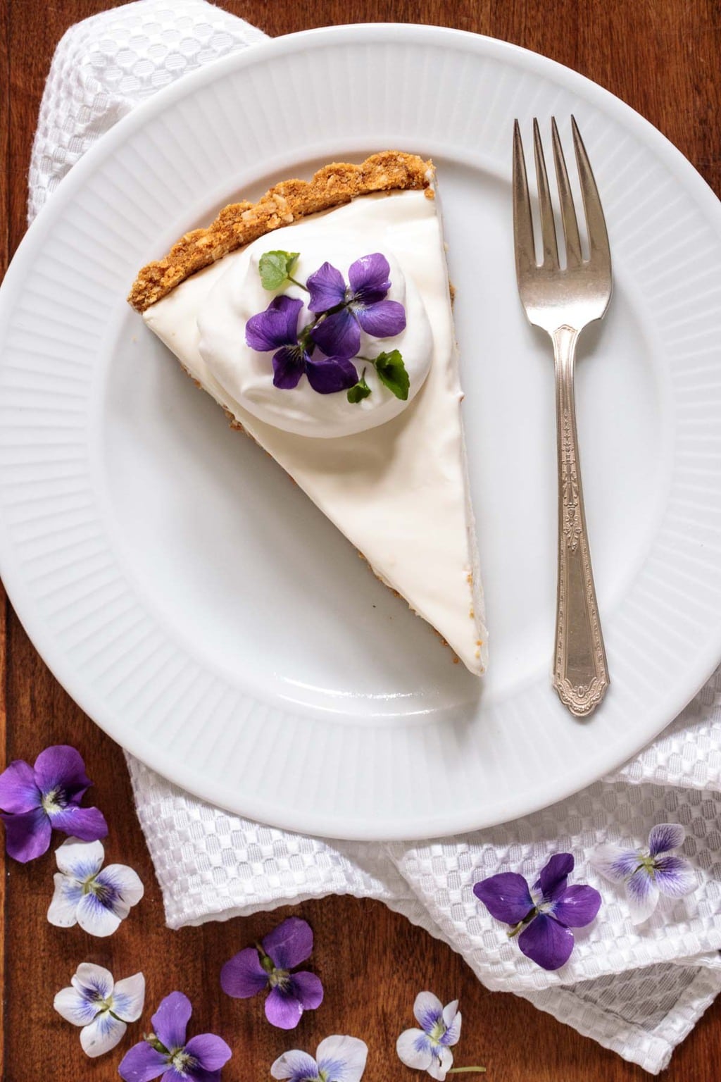 Overhead photo of a slice of Limoncello Lemon Tart on a white serving plate, surrounded by wild violets.