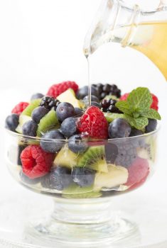 Vertical photo of Limoncello Syrup being poured over a bowl of fresh fruit.