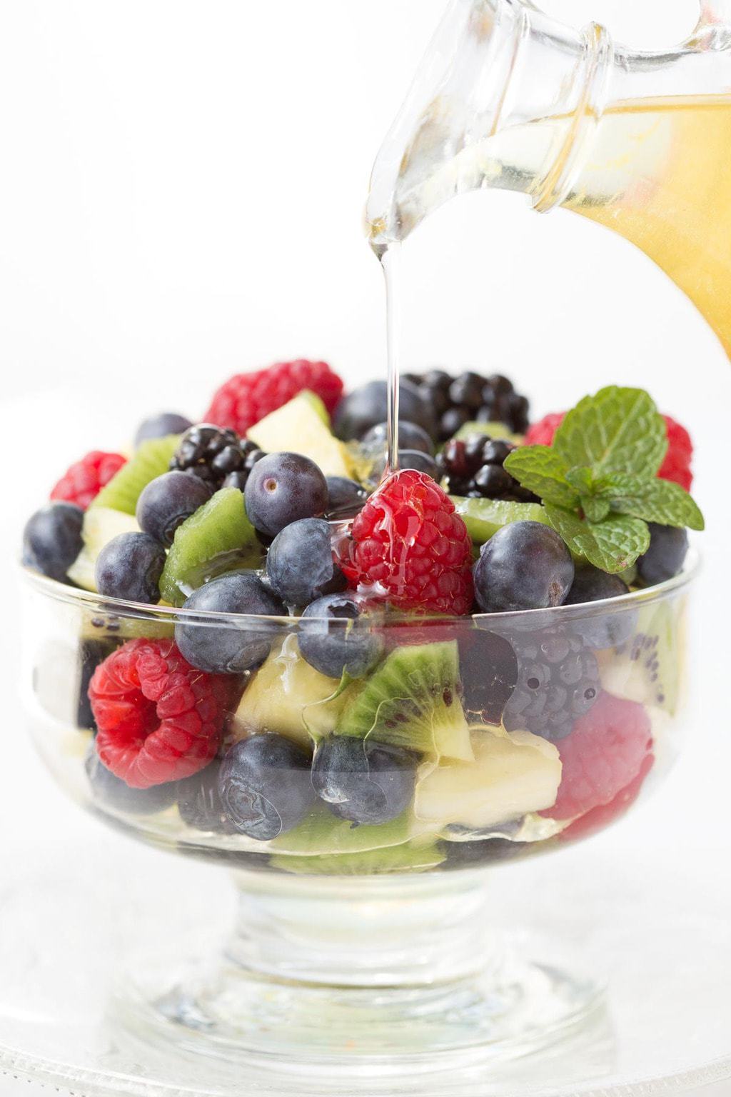 Vertical photo of a glass pitcher of Limoncello Syrup being poured over a bowl of fresh fruit.