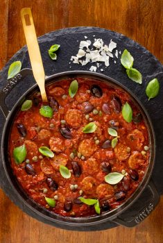 Vertical overhead photo of a Staub pot of Italian Sausage Puttanesca on a round slate surface.
