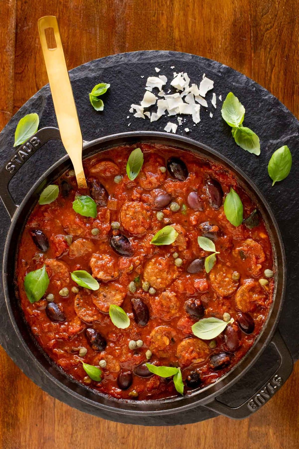 Vertical overhead photo of a Staub pot of Italian Sausage Puttanesca on a round slate surface garnished with fresh basil leaves.