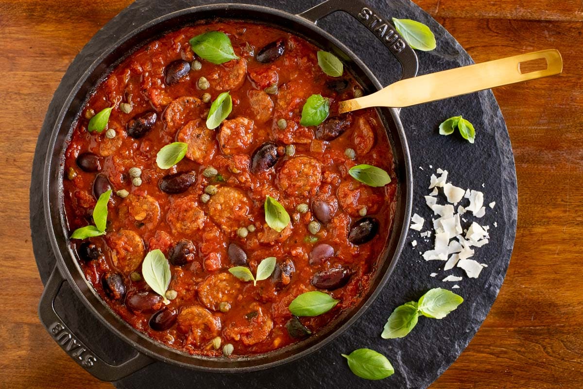 Overhead vertical photo of a Staub pot filled with Italian Sausage Puttanesca on a round slate stone.