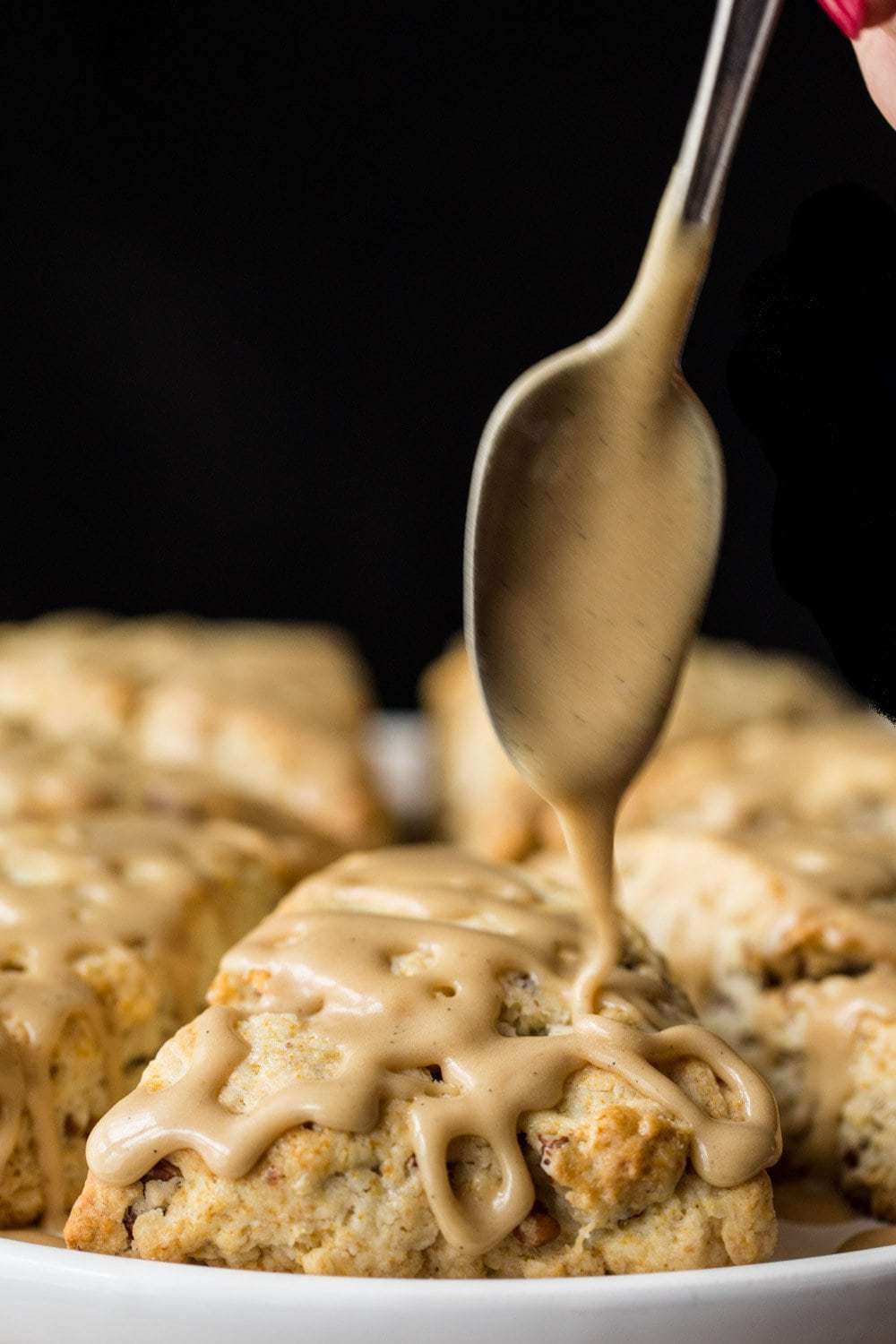 Closeup vertical photo of Ridiculously Easy Maple Pecan Scones on a platter with a spoon drizzling glaze over top.