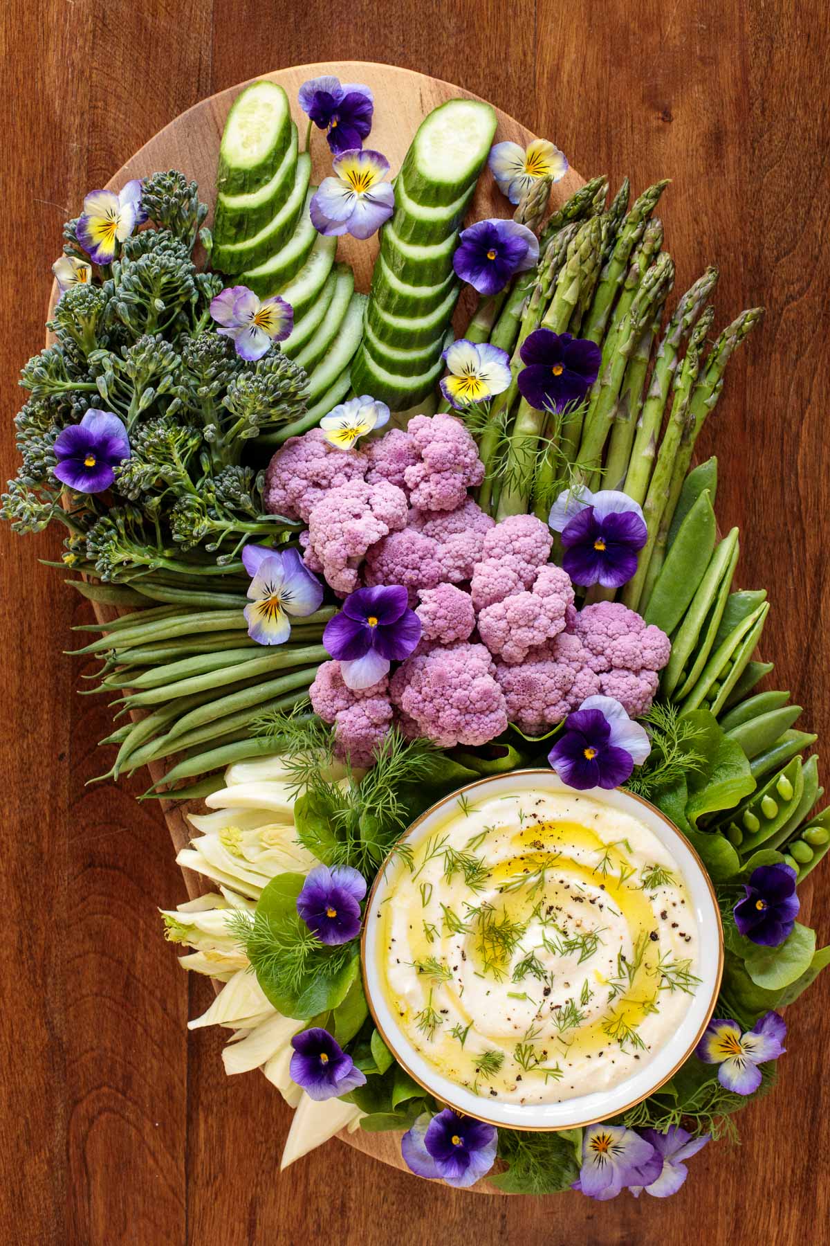 Vertical overhead photo of a veggie platter featuring Mediterranean Lemon Feta Dip on an oval wood appetizer platter decorated with pansies.
