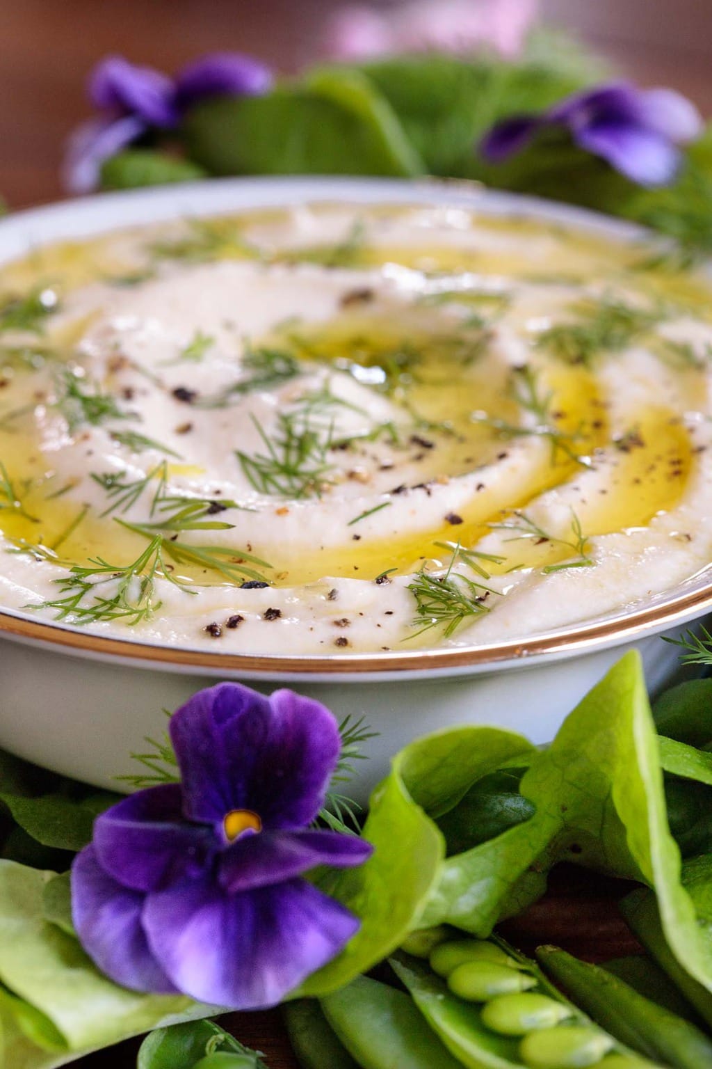Closeup photo of a white bowl of Mediterranean Lemon Feta Dip surrounded by vegetables and decorative edible flowers.
