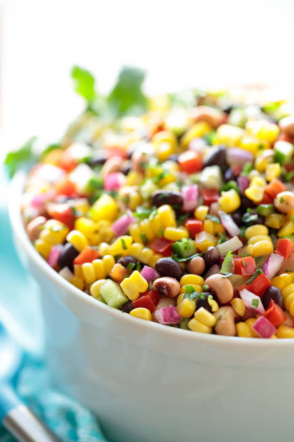 Vertical close up photo of Mexican Corn and Bean Salad in a white serving bowl surrounded by a turquoise napkin.