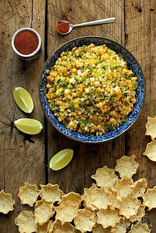 Vertical overhead photo of Mexican Street Corn in a blue and white bowl surrounded by chips and lime wedges.