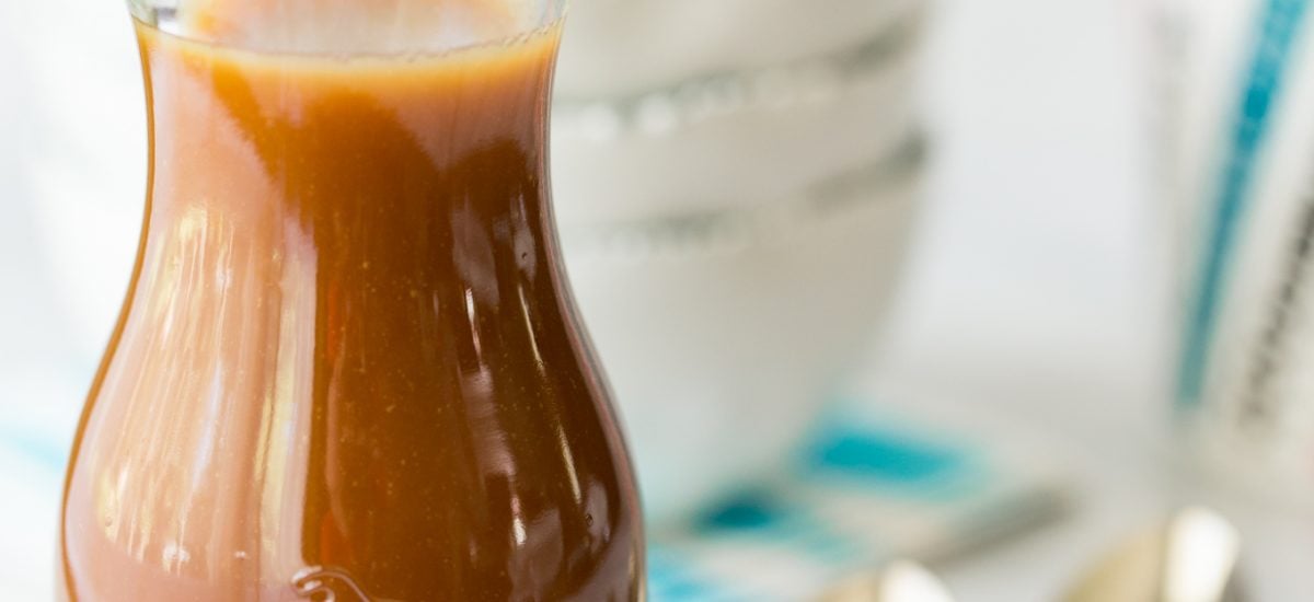Vertical picture of caramel sauce in a glass jar with small bowls and spoons in the background