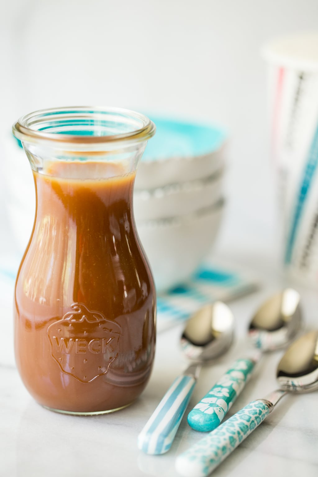 Vertical picture of caramel sauce in a glass jar with small bowls and spoons in the background