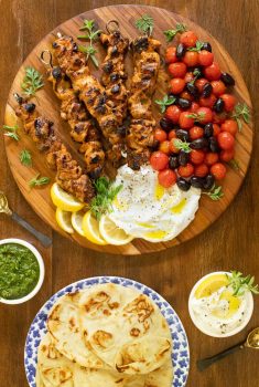Vertical overhead photo of Middle Eastern Chicken with Toum on a wood table with condiments in bowls nearby.