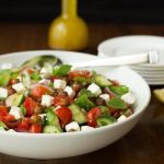 Horizontal image of Moroccan Tomato Cucumber Salad with Crispy Chickpeas in a white bowl with dressing behind it.