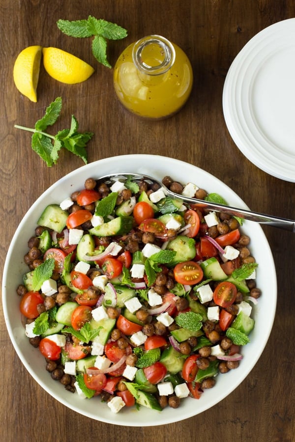 Overhead photo of a Moroccan Tomato Cucumber Salad with Crispy Chickpeas in a white bowl with the dressing in a cruet and sliced lemons and mint surrounding the plate.