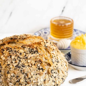 A loaf of No-Knead Seeded Oatmeal Bread with honey and butter on a white background.