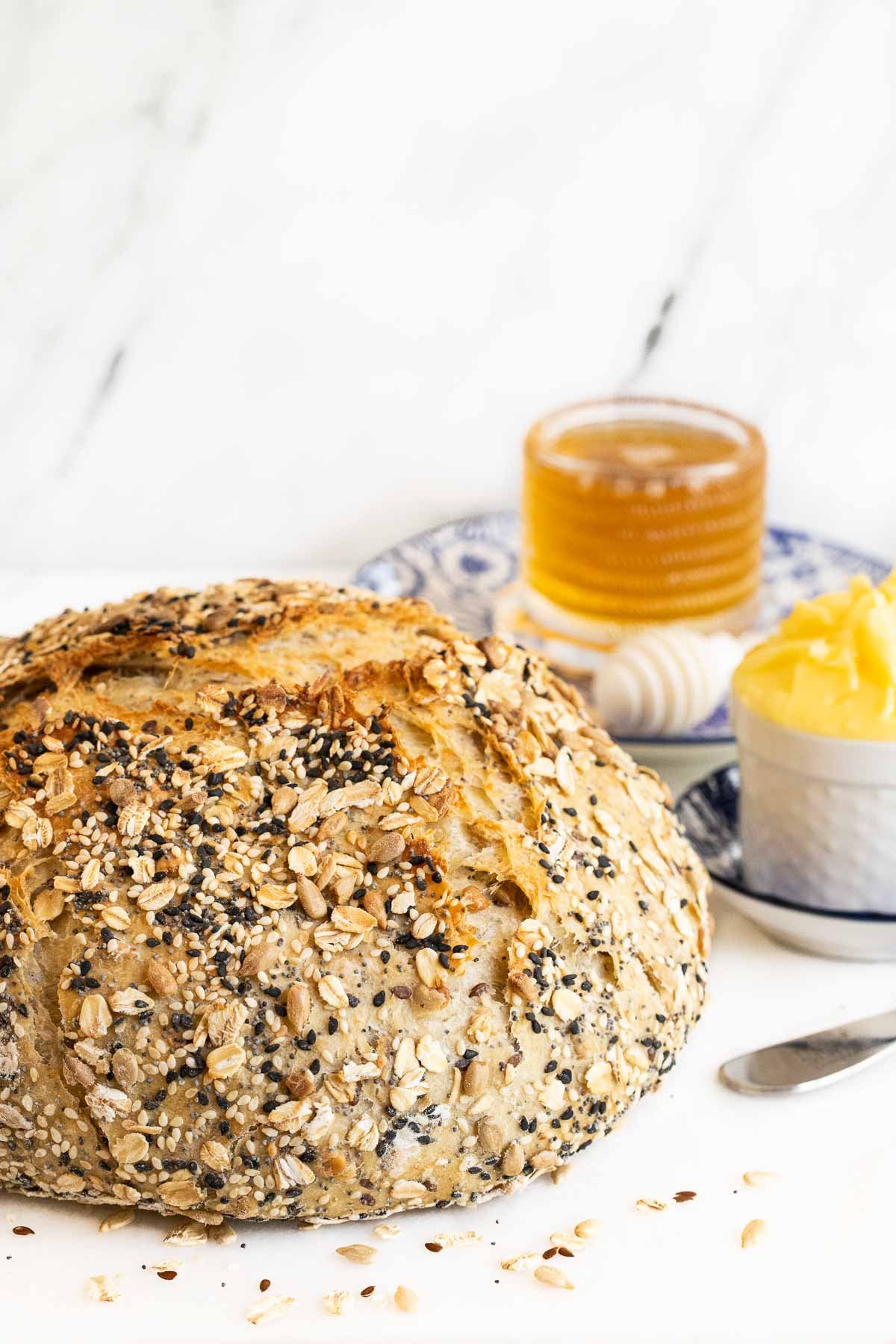 Vertical closeup photo of a loaf of No-Knead Seeded Oatmeal Bread with cups of honey and butter on a white marble background.