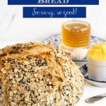 Closeup vertical photo of a loaf of No-Knead Seeded Oatmeal Bread with glass jars of honey and Irish butter in the background.