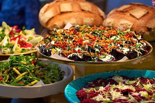 Photo of large platters of lunch vegetable entrees with loaves of bread in the background at Nopi in London, England.