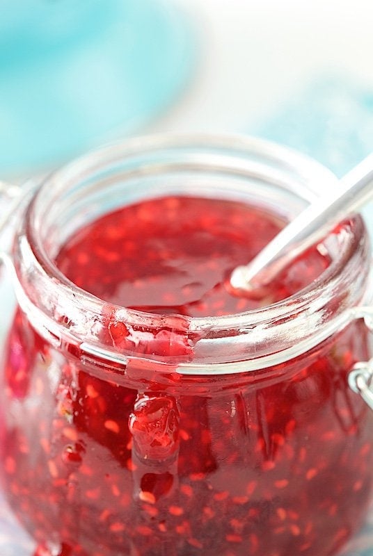 Closeup photo of the side of a glass jar of Old Fashioned Raspberry Preserves with a spoon in the jar and preserves dripping down the outside of the jar.