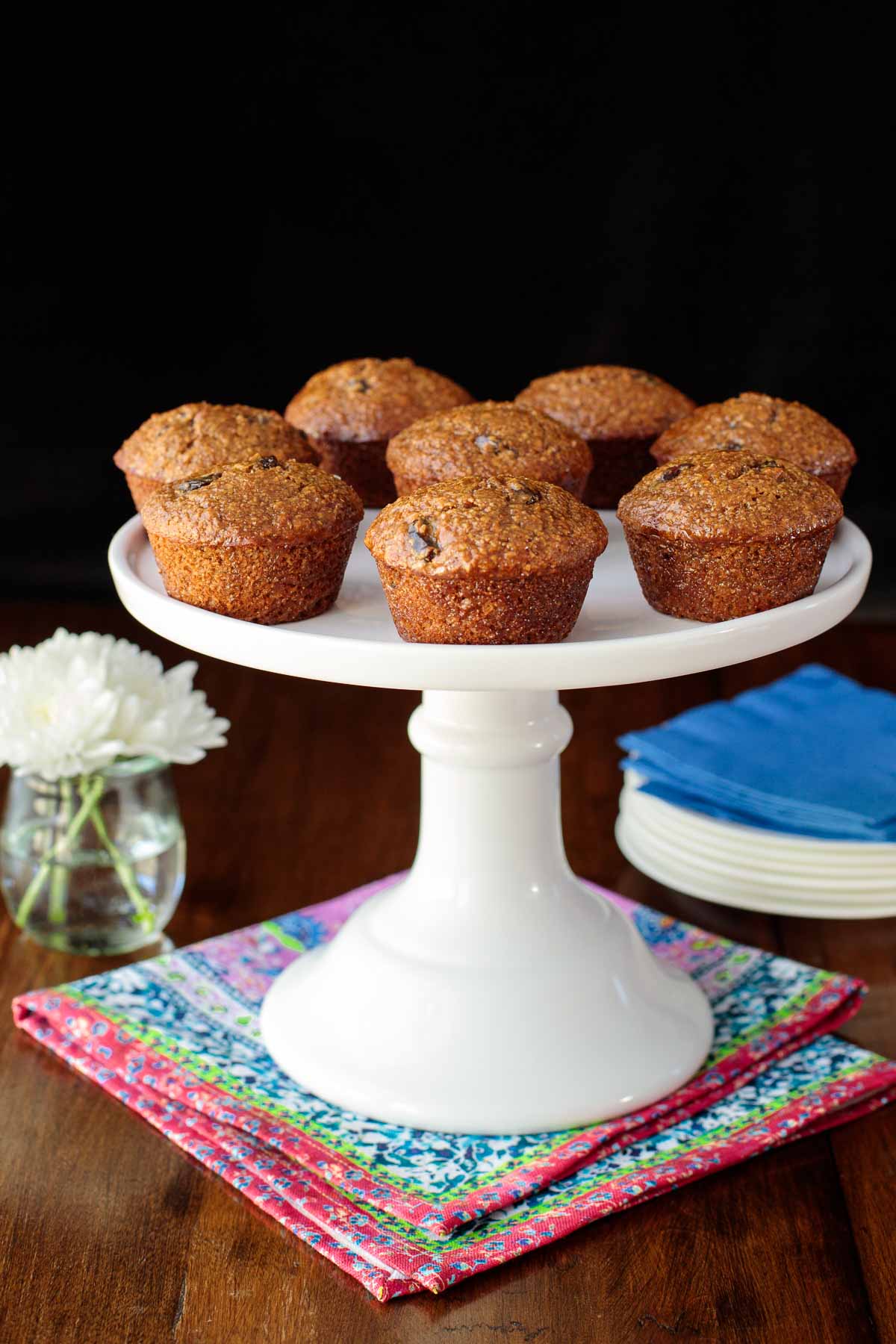 Vertical photo of One Bowl Buttermilk Bran Muffins on a white pedestal cake stand on a wood table.