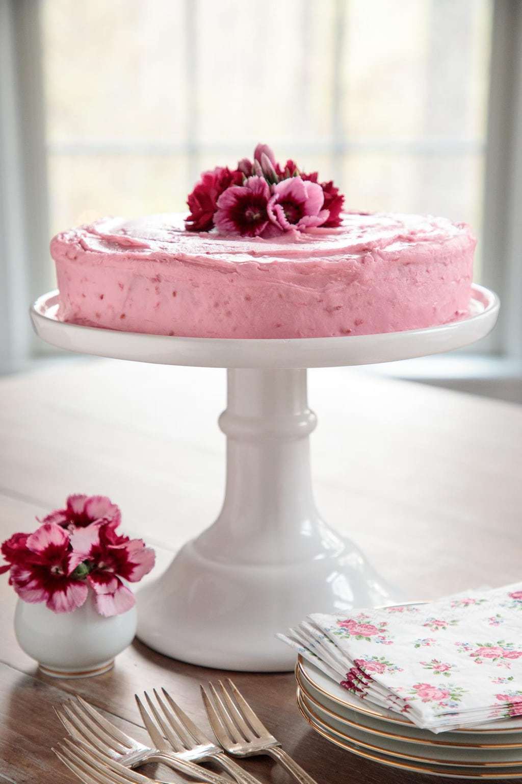 Vertical photo of a One-Bowl Buttermilk Cake with Fresh Raspberry Buttercream on a white pedestal cake stand with serving utensils and plates in the foreground and a window in the background.