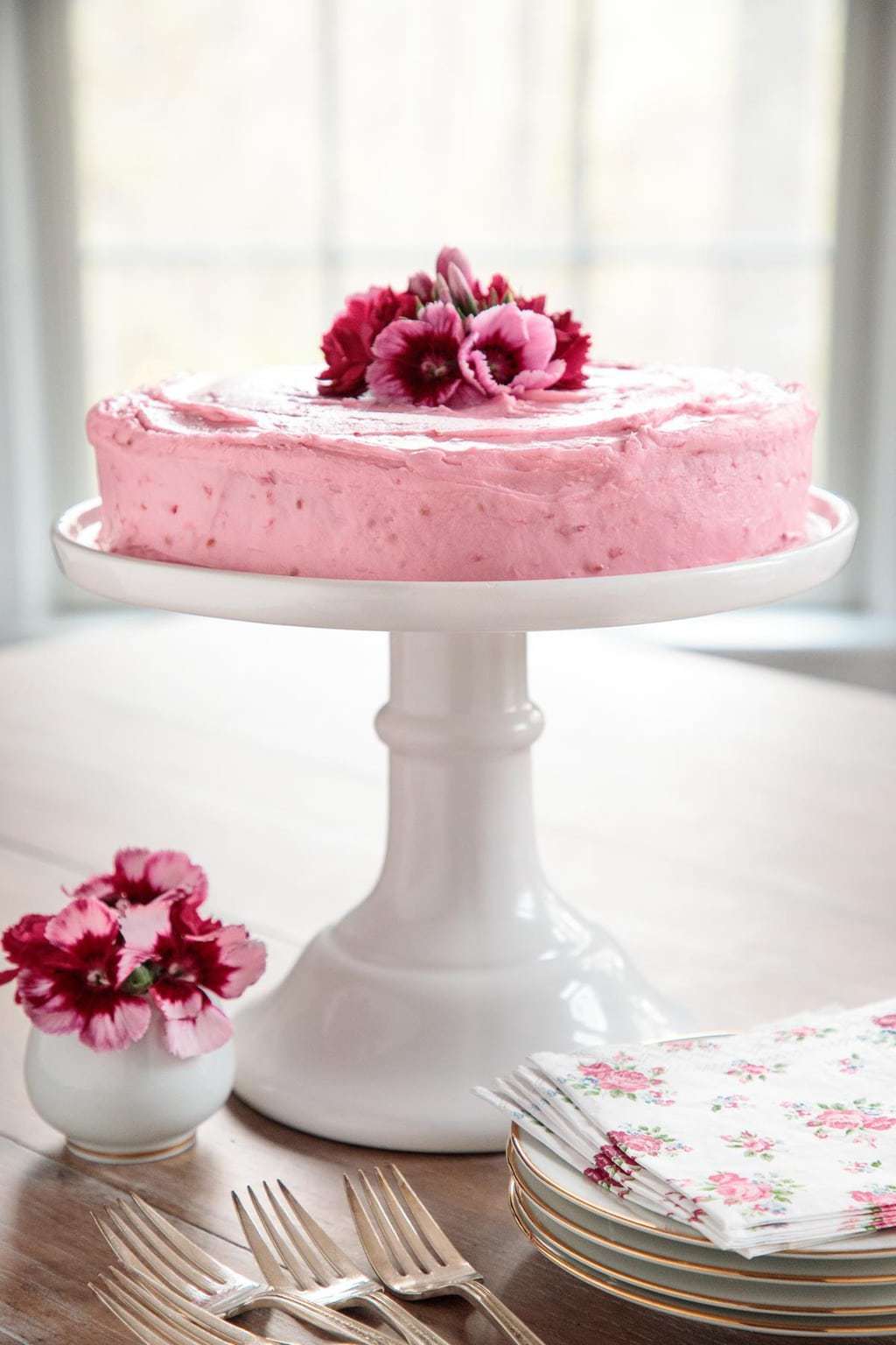 Photo of a One-Bowl Buttermilk Cake with Raspberry Buttercream on a white pedestal cake stand with serving plates, forks, napkins and flowers in the foreground.