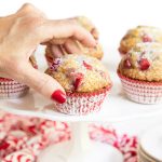 Closeup vertical photo of a hand reaching for a Fresh Strawberry Buttermilk Muffin on a white pedestal cake plate with other muffins in the background.