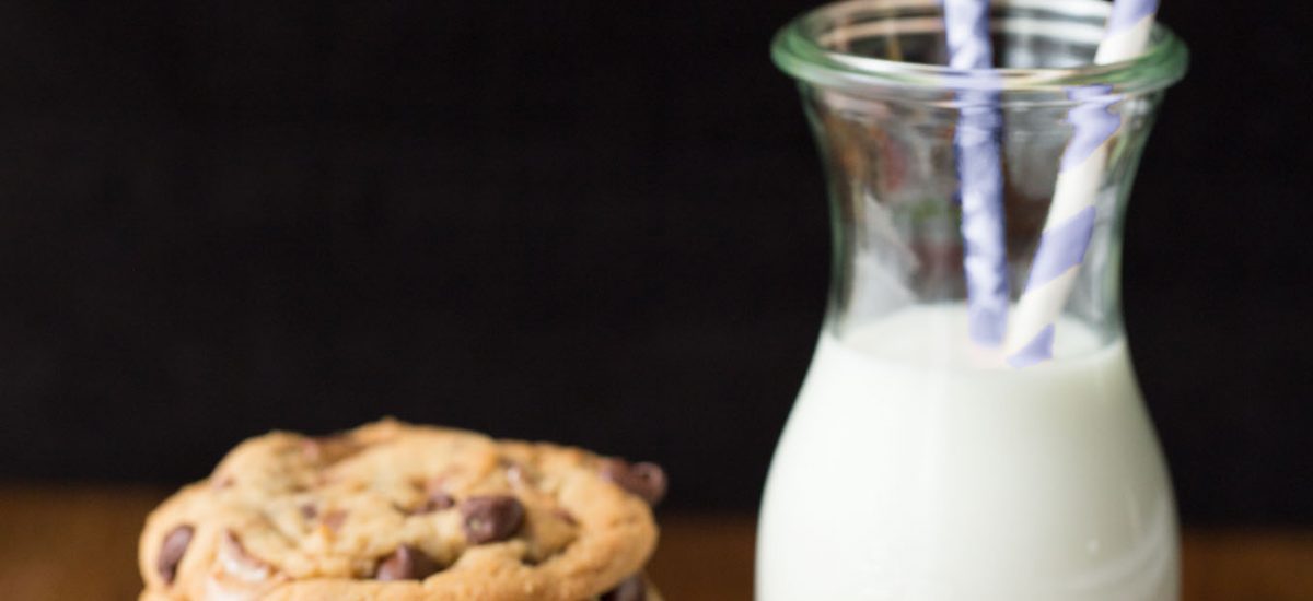 Vertical picture of Toffee Bar Chocolate Chip cookies stacked on a platter with a glass of milk and straws