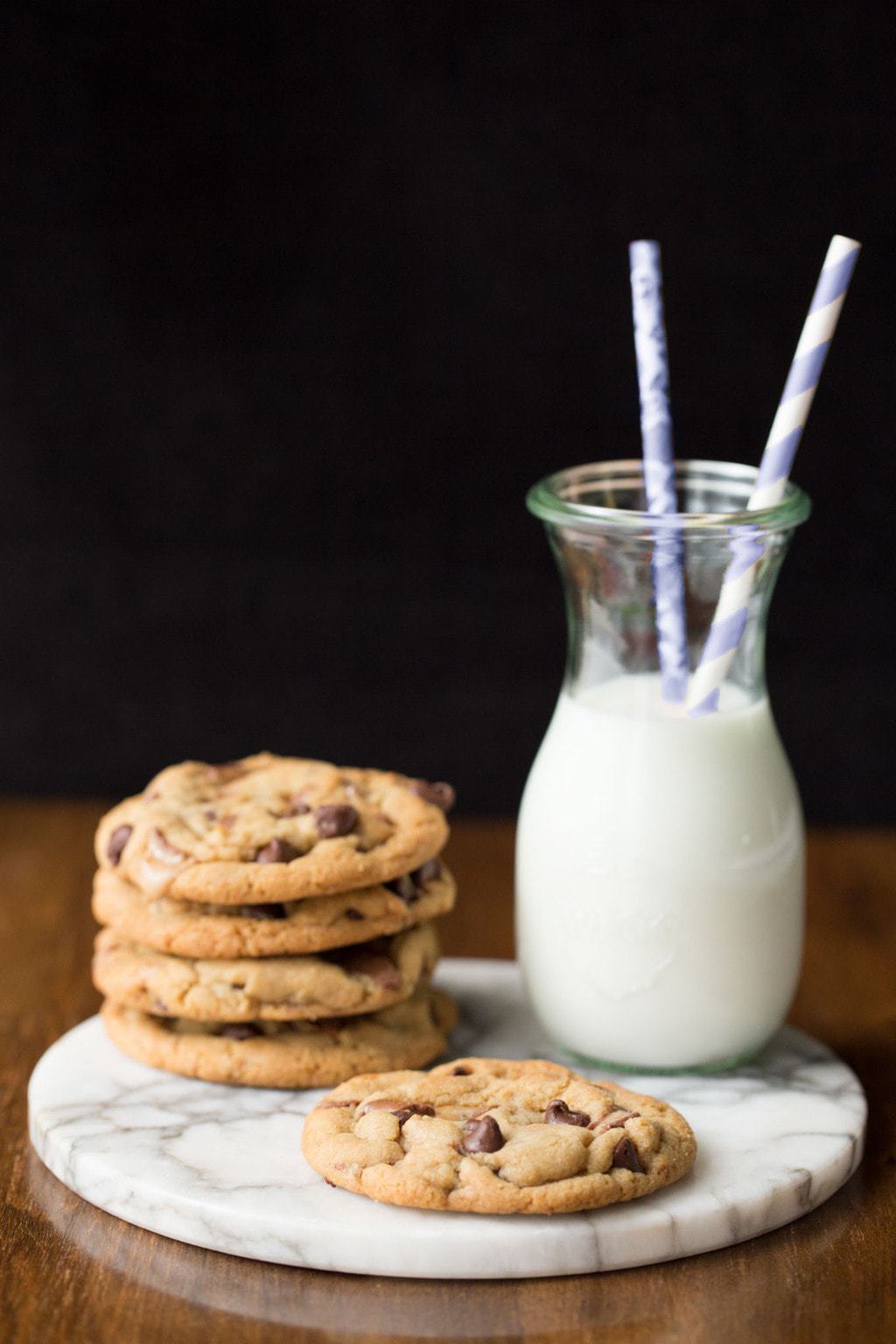 Vertical photo of Toffee Bar Chocolate Chip Cookies stacked on a marble platter with a glass of milk and straws.