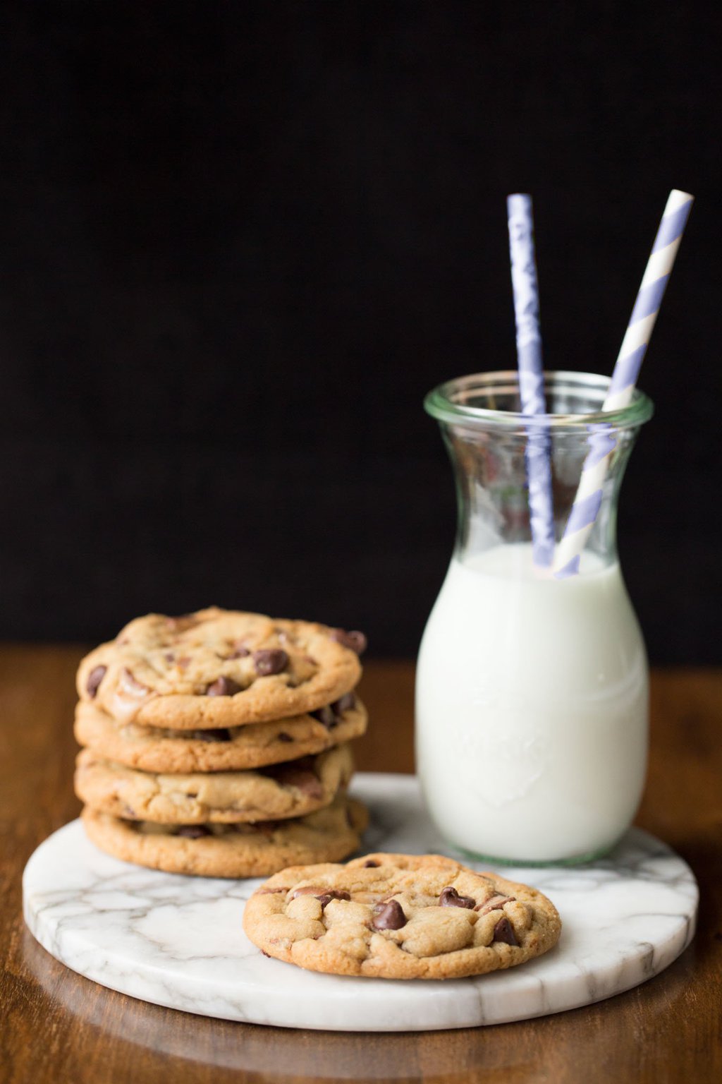 Vertical photo of a stack of One Bowl Toffee Bar Chocolate Chip Cookies on a round marble slab. A carafe of milk with two straws is next to the stack.
