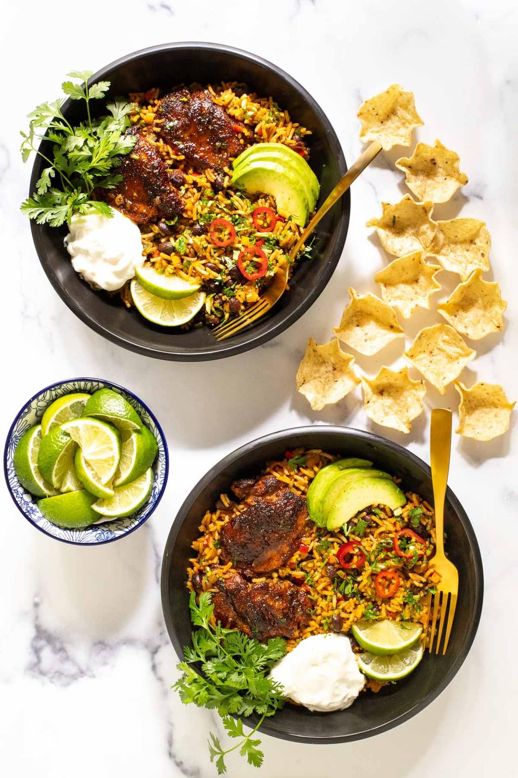 Overhead vertical photo of One-Pot Mexican Chicken and Rice in black serving bowls with tortilla chips and lime wedges.