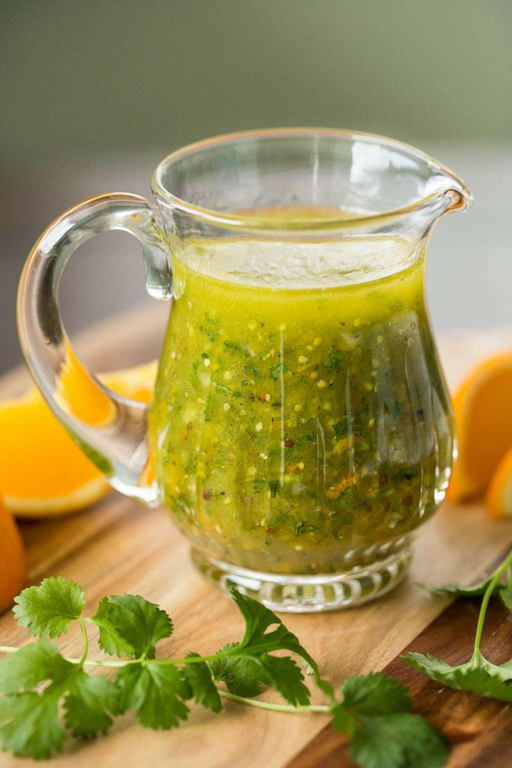 Photo of a glass pitcher filled with Orange Cilantro Vinaigrette on a wood cutting board surrounded by orange wedges and cilantro leaves.