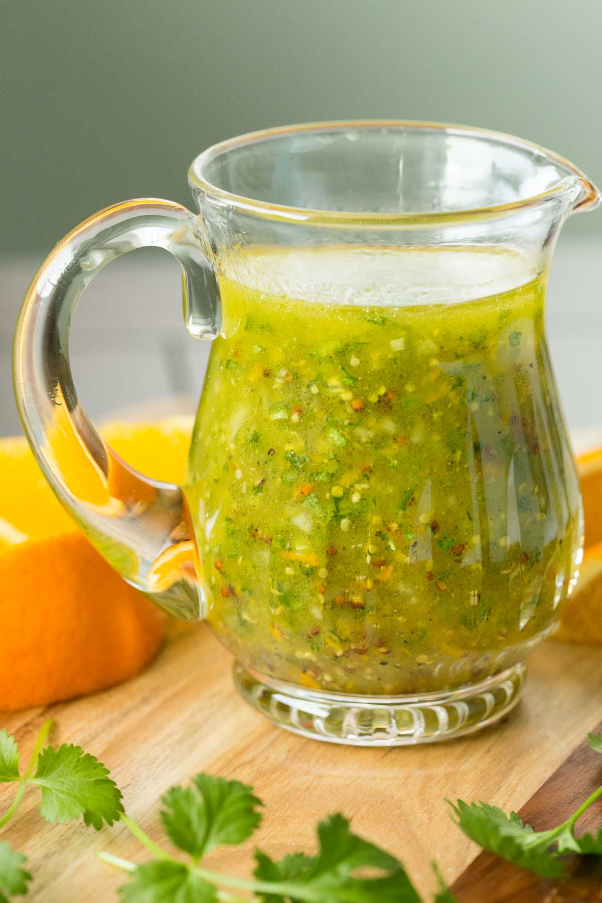 Photo of a glass pitcher filled with Orange Cilantro Vinaigrette on a wood cutting board surrounded by cilantro leaves and orange wedges.